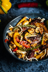 Wok with noodles, peppers, mushrooms, turkey meat and different vegetables and sesame seeds in a blue bowl on dark background