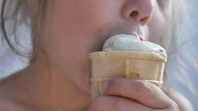 Small girl with plait eats tasty vanilla ice-cream in waffle glass and bites hungily with dirty mouth and looks closeup side view