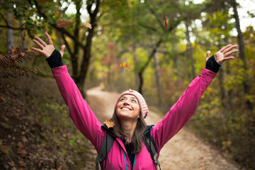 Hiking girl in pink on a trail in the forest. Hands up enjoying the falling leaves in nature in...