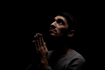 Headshot of serious confident young man praying in dark room. Lit from above and looking up
