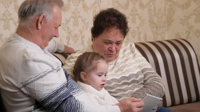 Happy family, modern grandparents play with a child, a girl sitting with a tablet online at home. Child, girl shows something in tablet to grandparents. Elderly couple with granddaughter and gadget