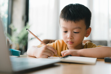 Asian Boy Doing homework with the intention. Child boy holding pencil writing, studying at home on a laptop to study from home during Covid-19 pandemic lockdown. Online education, Homeschooling.