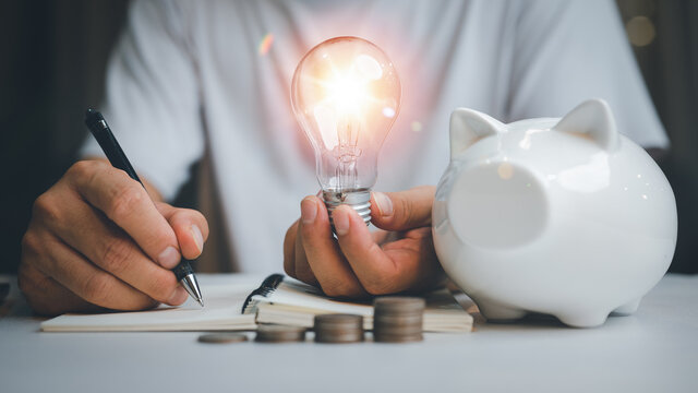 saving energy and money concept. idea for save or investment. businessman holding lightbulb on piggy bank and coins stacking on desk with note for planning money finance.