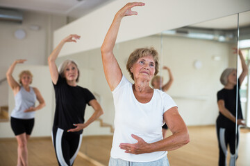 Fototapeta na wymiar Positive elderly woman practicing ballet dance moves during group class in choreographic studio.