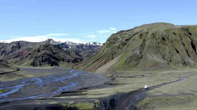 Aerial view of a 4x4 white car driving on a dirt road by a wide riverbed and range of green and grey mountains in the background in Iceland near Thakgil campsite