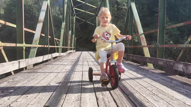 Cute blonde child riding tricycle on old wooden bridge - Sunlight passing through sidebeams of bridge - Handheld moving bakwards while filming girl cycling against camera - Eidslandet Norway