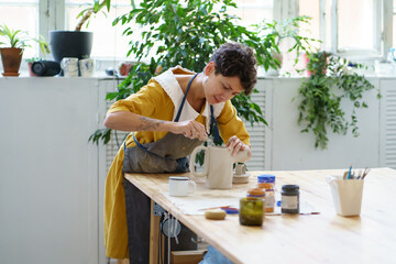 Young woman ceramist decorating potter jug after burning. Artisan female paining pottery in ceramic workshop studio. Craftswoman working on handicraft. Artistic hobby and entertainment leisure concept