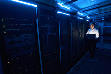 Male server technician walking with laptop in data center room