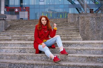 a young adult red-haired woman in a red jacket, blue jeans and red sneakers is sitting on the steps of a concrete staircase. There is a glass building in the background. Daylight, everyday lifestyle. - Powered by Adobe