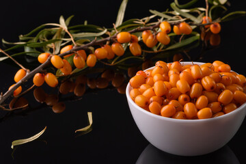 A sea buckthorn berry in a white cup stands on a black reflective surface in close-up. Nearby lies a branch of sea buckthorn.