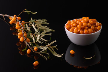 a branch of sea buckthorn and a white cup filled with sea buckthorn berries on a black reflective surface