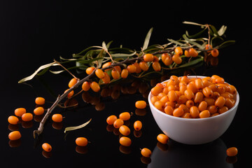 sea buckthorn berry in a white cup stands on a black reflective surface. A branch lies nearby and sea buckthorn berries are scattered.