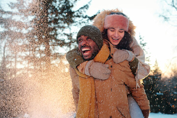 Cheerful multiracial couple in winterwear laughing while girl embracing her boyfriend during...