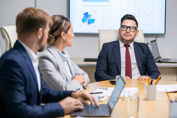 Confident business coach in suit and eyeglasses listening to one of young employees at conference
