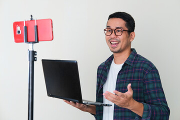 Adult Asian man holding laptop computer while presenting using mobile phone video call conference