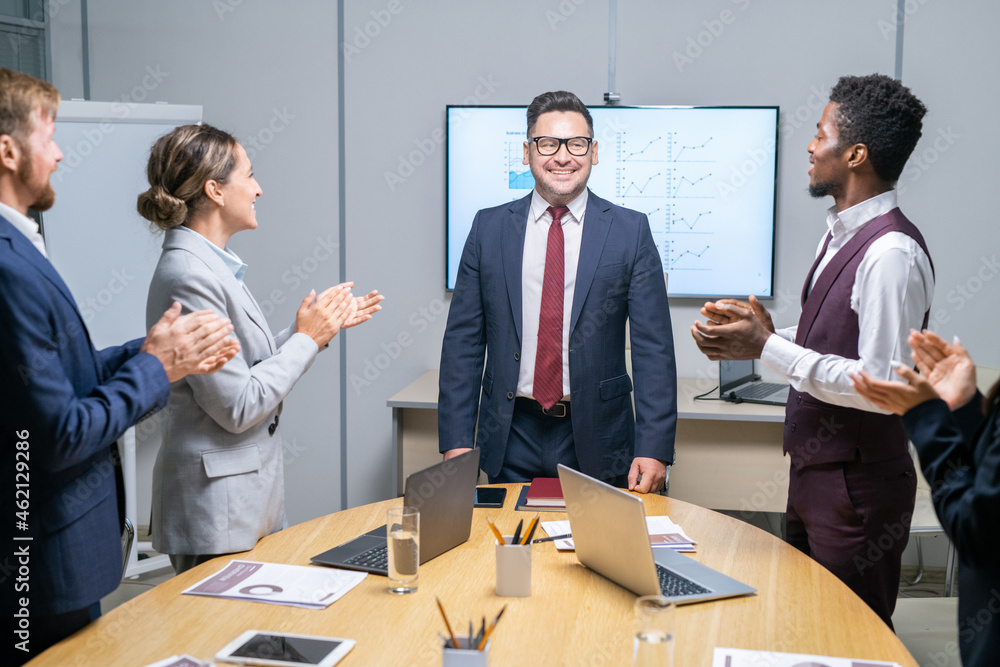 Poster intercultural employees standing by large table and clapping hands while congratulating their boss i