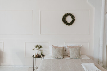 Modern bright home bedroom interior with bed, neutral bed linens, pillows, Christmas wreath made of...