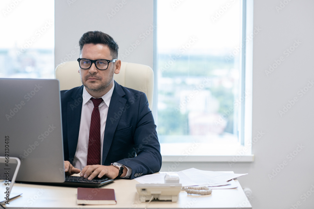 Poster mature businessman in eyeglasses and suit working in front of computer monitor by workplace
