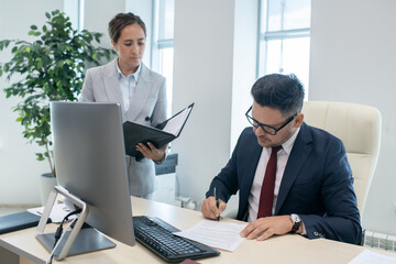 Young pretty secretary looking at her boss signing contract by workplace