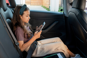 Side view of cute girl scrolling in smartphone while sitting on backseat of electric car