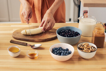 Hands of young female chopping banana on wooden board for homemade smoothie