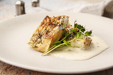 Plate with delicious baked cod fillet, sauce and greens on table in restaurant, closeup