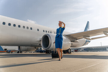 Smiling stewardess holding black suitcase and standing on runway with airplane in the background