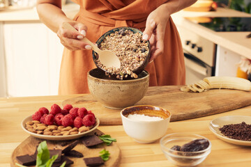 Hands of female with wooden spoon putting muesli into bowl while preparing dessert