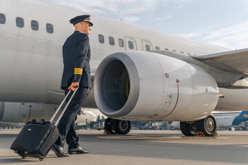 Back view of pilot in black suit walking with travel bag near plane. Profession concept - obrazy, fototapety, plakaty