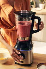 Young woman pressing button of electric blender while mixing and shaking smoothie ingredients by...