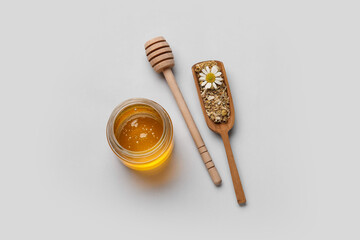 Jar of sweet honey and scoop with dried chamomile flowers on grey background