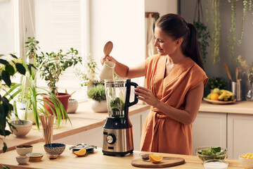 Happy young housewife pouring milk into electric blender while preparing homemade smoothie or...