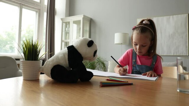 Smilling happy girl sitting in the table with a toy panda bear near to her enjoying creative activity, drawing pencils coloring pictures in albums.