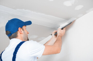 A master in uniform applies putty with a spatula to the reinforced joint of drywall on the ceiling.