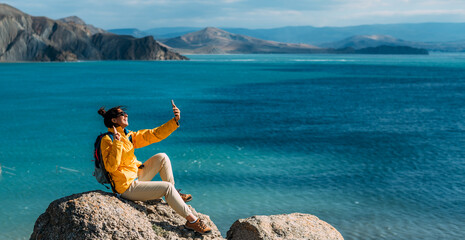 A female traveler is talking on a video call against the background of the sea, panorama. A traveler takes a selfie on her smartphone against the background of the sea. A traveler on the seashore.