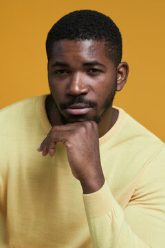 Vertical portrait of handsome African-American man looking at camera while posing resting chin on hand against yellow background in studio