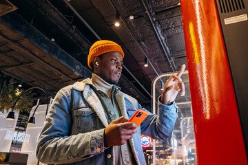 Smiling African-American man in warm denim jacket with wireless earphones uses self-service kiosk...