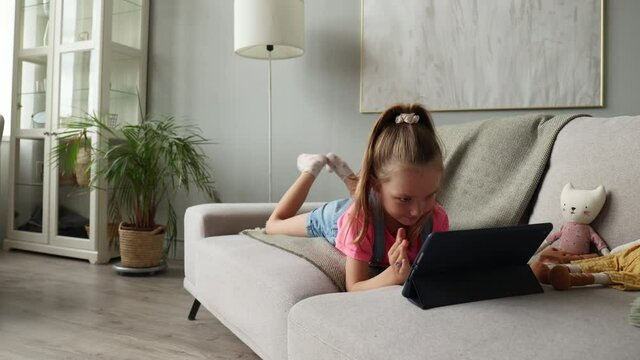 Smiling little girl lying on sofa playing online games, web surfing information, using funny applications on tablet