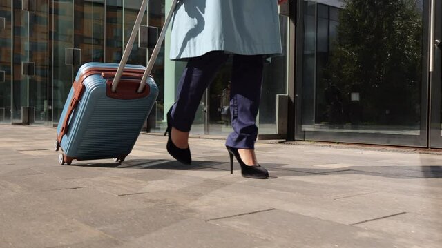 Closeup view of young woman with suitcase in hands walking through city in autumn spbi. Shot of feet of businesswoman holds luggage in hand and walks actively on street along glass building on sunny
