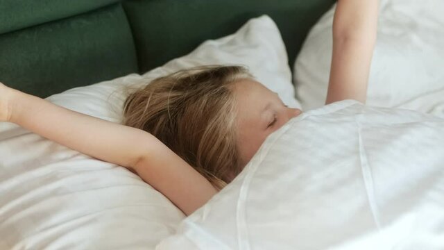 Cute little girl child wakes up in the morning in bed in the bedroom on an orthopedic mattress and pillow after a healthy sleep and does not want to wake up and hides under the covers.