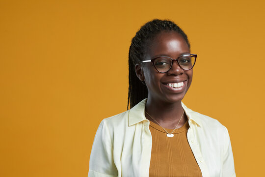 Portrait of young African-American woman wearing glasses and smiling at camera while posing against yellow background in studio, copy space