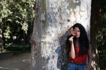 a beautiful, dark-haired, curly-haired adult woman with latin features is leaning against a giant tree. The woman covers her eye with a dry leaf that falls from the tree in autumn. Autumn concept