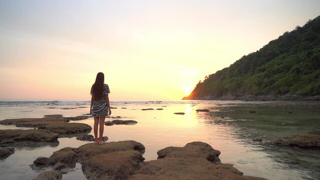 A young woman with her back to the camera stands on the exposed rocks from the low tide as she admires the colorful sun setting behind a mountain. Title space