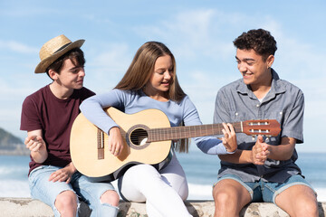 Group of happy multiethnic teenagers singing and playing the guitar sitting at the seaside.