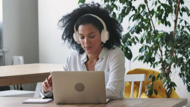 Portrait of African American woman talking on video conference call using laptop and headphones taking notes on notepad. Brunette sits at table in home office. Close up. Slow motion ready 59.97fps.