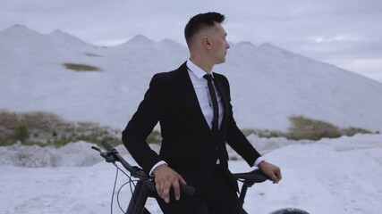 Serious respectable male entrepreneur in elegant black suit and tie standing with crossed arms and leaning on parked bike in highlands while looking away with determination and aspiration 