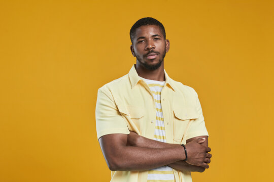 Waist up portrait of handsome African-American man looking at camera while posing confidently with arms crossed against yellow background in studio, copy space