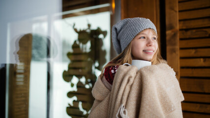 Portrait of preteen girl wrapped in blanket with hoit drink outdoors in front of wooden cottage in winter nature.