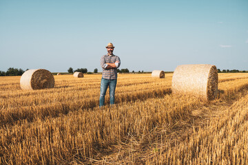 Naklejka na ściany i meble Happy farmer is standing beside bales of hay. He is satisfied because of successful harvesting.