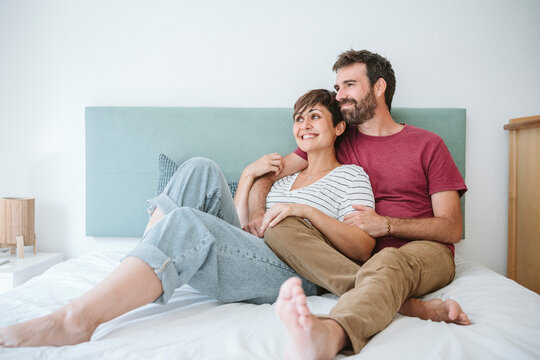 Happy mid adult couple looking away while sitting together on bed at home